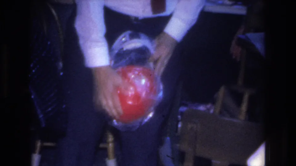 Final 8mm frame of a man in white dress shirt holding a red cellophane-wrapped sphere at crotch level, with a second party guest partially visible at right, 1966 Hoboken interior.