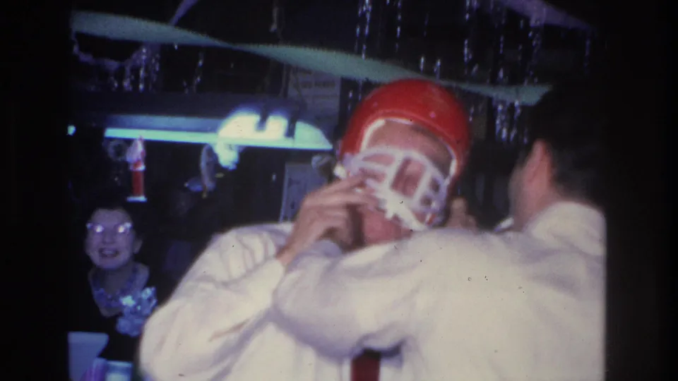 Man in white dress shirt and red tie adjusting a small red football helmet at a party; female guest with glasses laughs behind him; Hoboken, NJ, 1966.