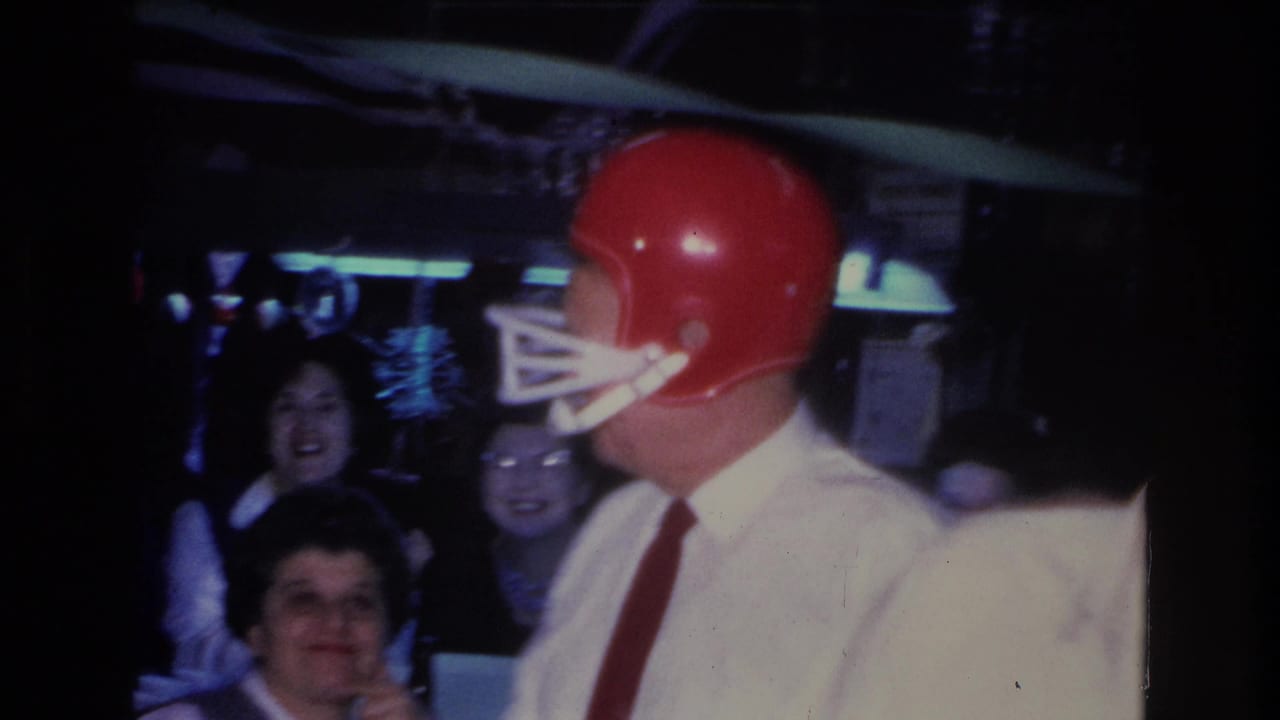 Man in white shirt and red tie wearing red football helmet shown in profile; two female guests smile in background; blue neon lighting; Hoboken, NJ, 1966.