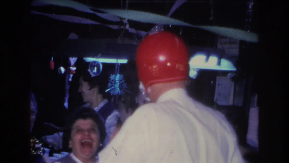 Rear view of man in white shirt wearing red football helmet; a woman laughs openly in the foreground; tinsel streamers and a sunburst ornament hang in background; Hoboken, NJ, 1966.