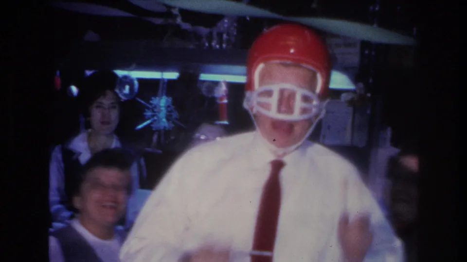 Man in white shirt and red tie wearing red football helmet faces camera with drink in hand; a young figure and other guests visible behind him; Hoboken, NJ, 1966.