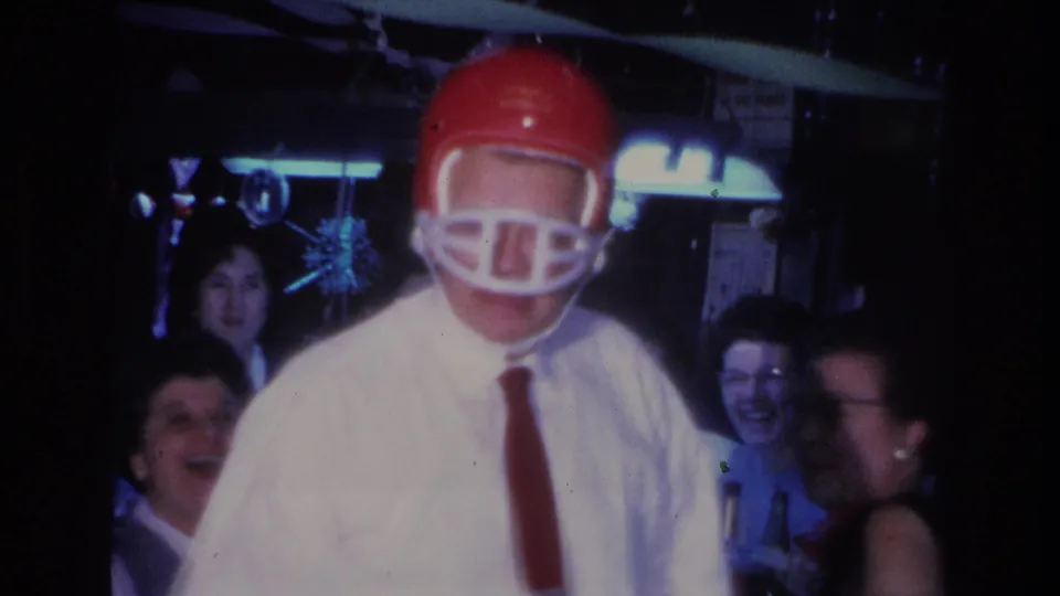 Man wearing a red football helmet as a party novelty at a 1966 indoor gathering in Hoboken, New Jersey; laughing female guests and sparkly hanging decorations visible in the background.