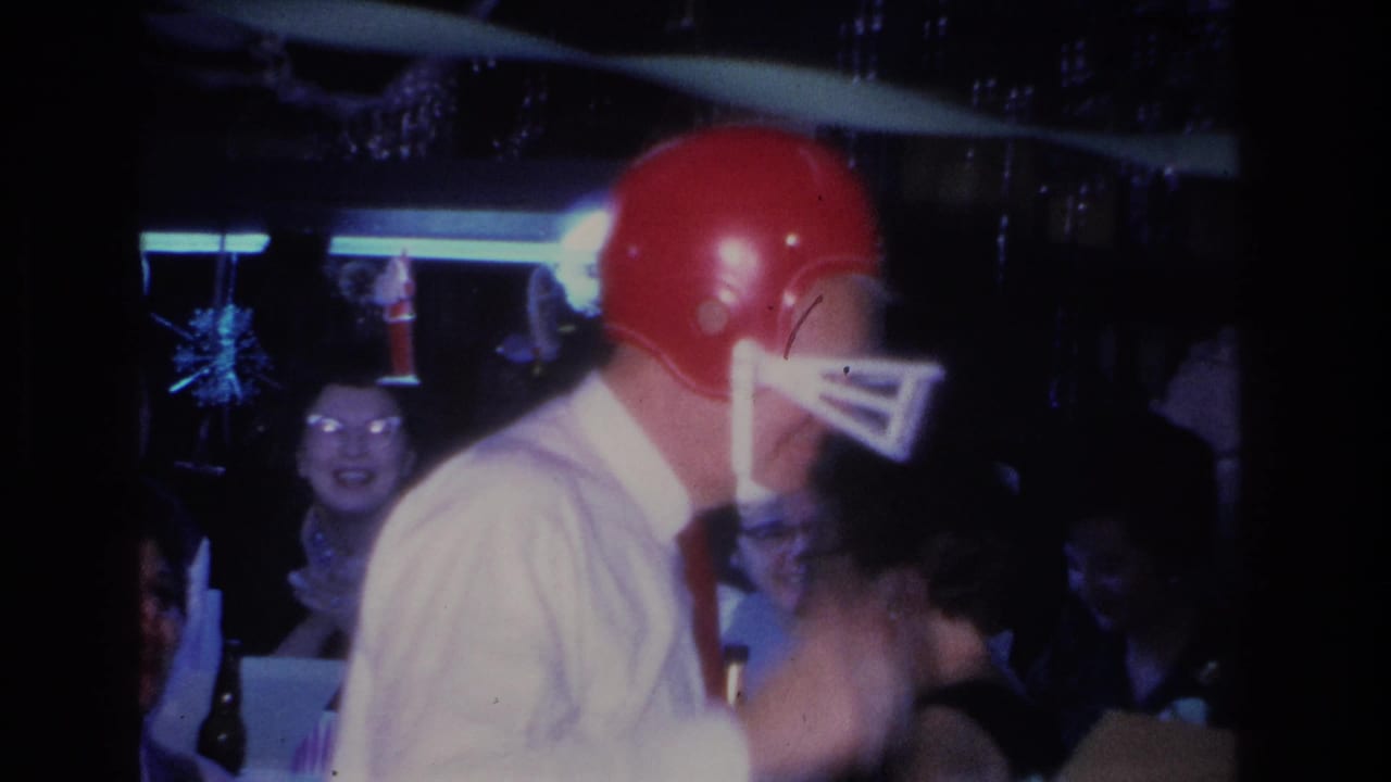 Man in white shirt and red football helmet seen from the side at a dim indoor party, with a smiling female guest and hanging decorations visible behind him.
