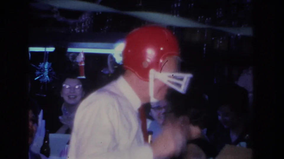Side profile of man in red football helmet at a 1966 indoor gathering in Hoboken, New Jersey; a female guest with glasses smiles in the background near sparkly blue hanging decorations.