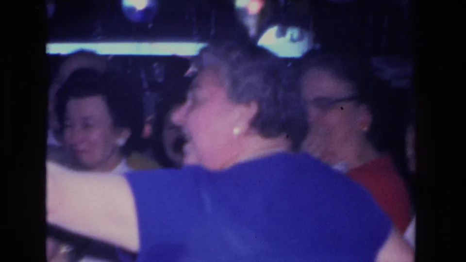 Close-up of adult guests at a 1960s indoor party; woman with cat-eye glasses and short gray hair visible mid-frame amid dim mixed-color ambient lighting.
