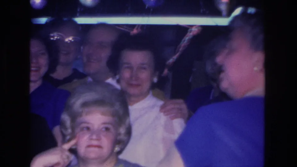 Group of adult guests at a 1960s indoor gathering in Hoboken, New Jersey; a woman in the foreground gestures near her face while others smile around her in dim blue-tinted lighting.