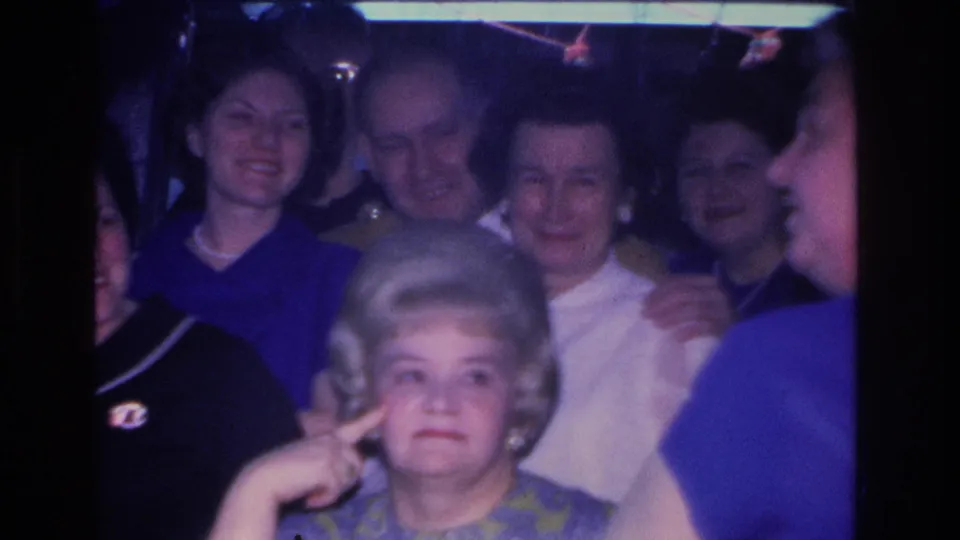 1966 Hoboken, New Jersey indoor party: a woman with a bouffant hairstyle makes a playful gesture amid a crowd of adults in 1960s attire; 8mm film color shift visible.