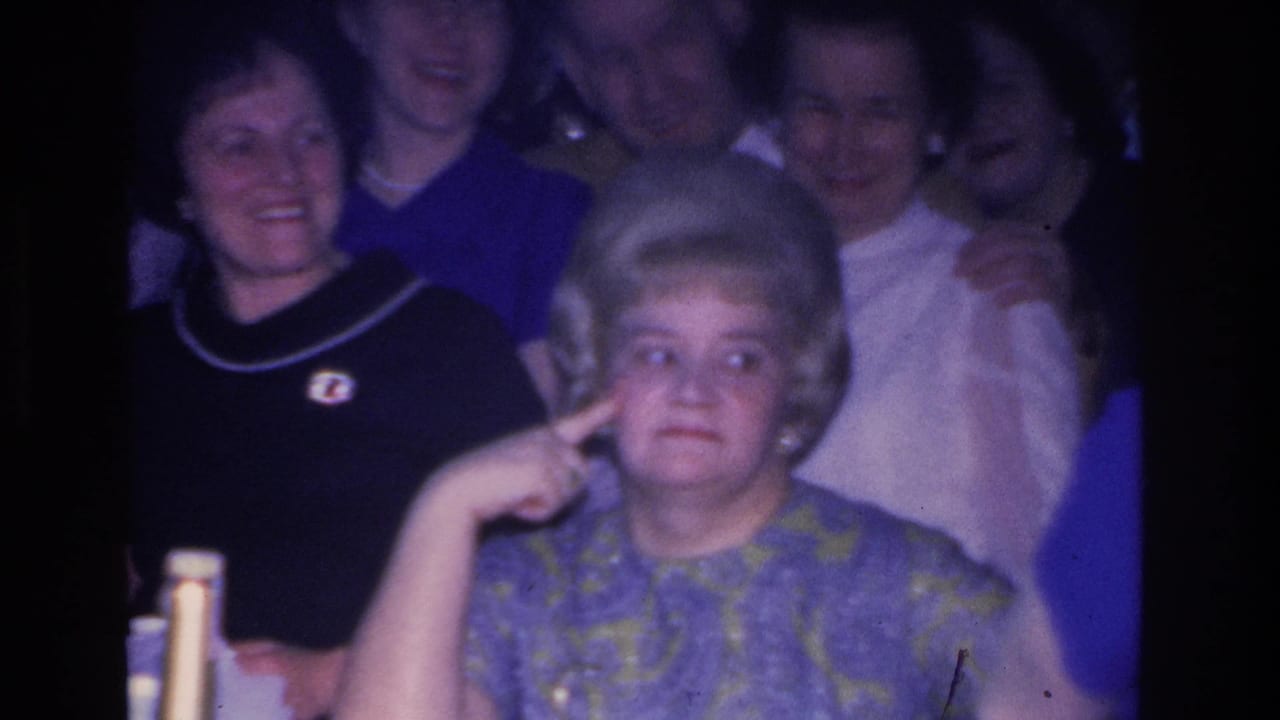 1966 indoor party scene: close frame of a woman with set bouffant hair mid-gesture, with another woman in a navy top and brooch smiling behind; vintage 8mm blue cast present.
