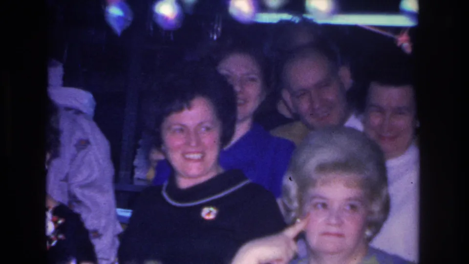 1966 Hoboken party, wider shot: colorful hanging bulb string lights overhead, a smiling woman in a navy top with brooch, and the platinum-haired woman mid-gesture among adult guests.