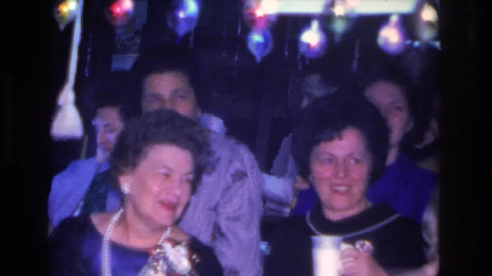 1966 indoor party: a woman in a gray jacket and pearl necklace laughs alongside a woman in a dark top; vintage bulb party lights hang overhead and guests fill the background.