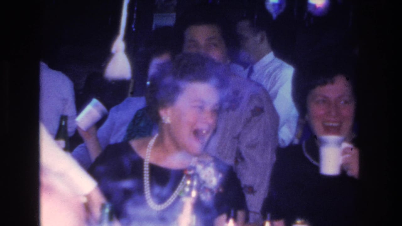 8mm home movie frame showing a woman in dark dress and pearl necklace laughing at an indoor party, consistent with 1966 Hoboken, New Jersey.