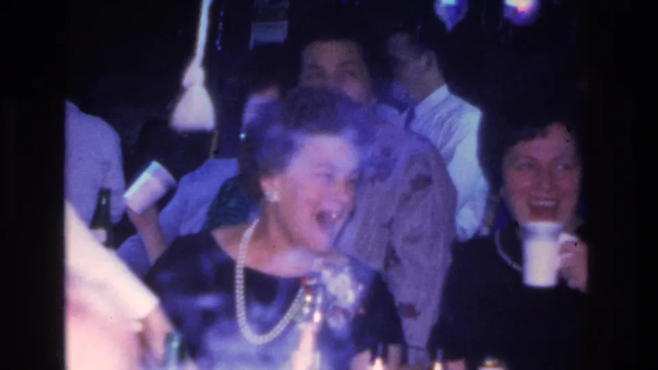 8mm home movie frame showing a woman in dark dress and pearl necklace laughing at an indoor party, consistent with 1966 Hoboken, New Jersey.