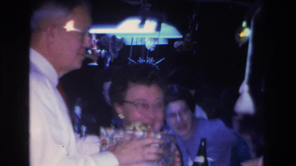 1960s 8mm home movie frame of a man in a white shirt and a woman in eyeglasses at a party table, with a hanging ceiling lamp and string lights in a dim interior.