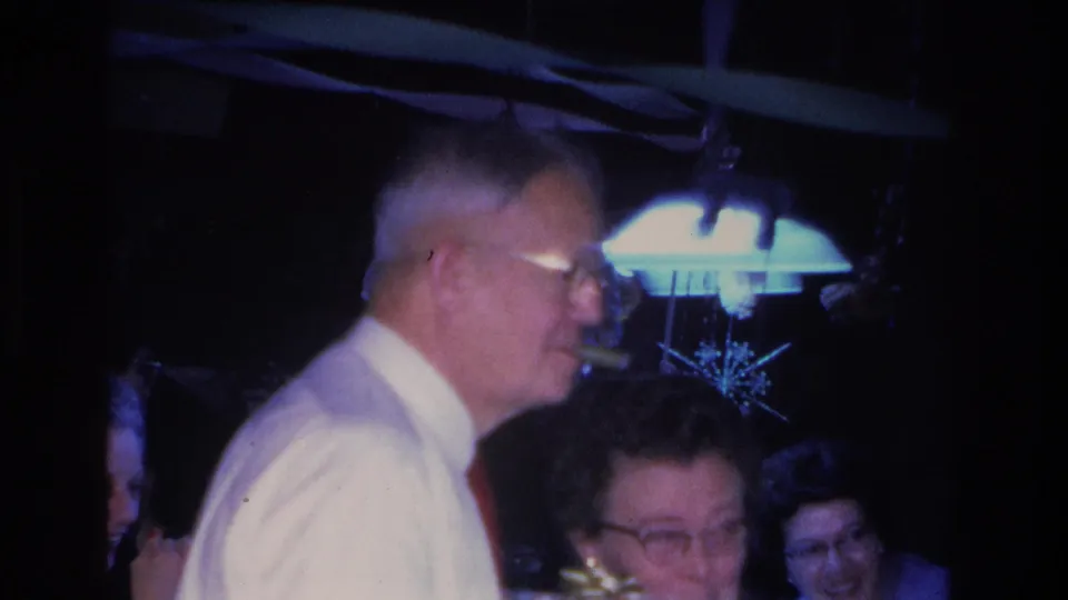 1960s 8mm home movie frame of a man in a white shirt at an indoor party, with a star-shaped decoration and hanging lamp visible in the dim background.
