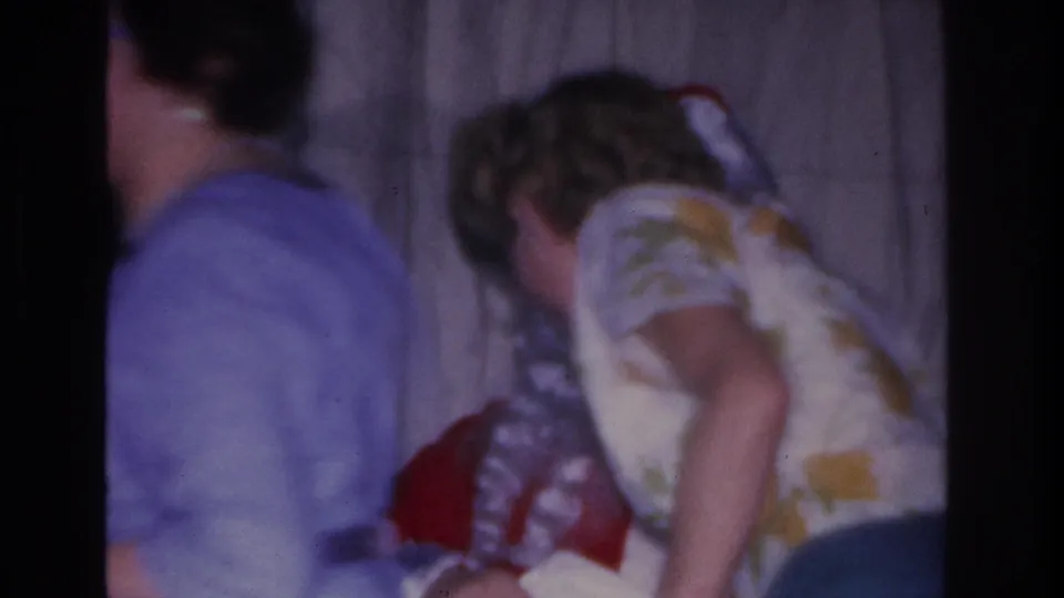 Two adults at a 1966 Hoboken, NJ Christmas gathering; person in floral blouse reaches toward a red bag, shot on 8mm film.