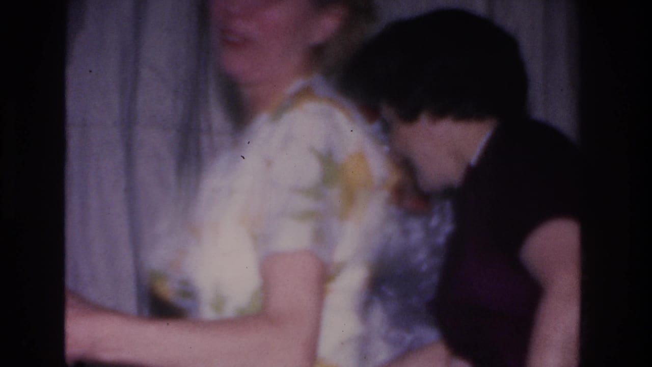 Two adults at a 1966 Hoboken, NJ Christmas gathering; floral-blouse and dark-top figures lean close together near a light curtain, 8mm film.