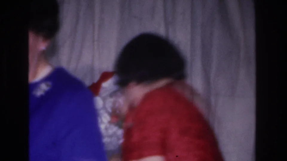 Woman in blue sweater leaning toward Santa, second person in red top visible behind, 8mm home movie, mid-1960s holiday scene