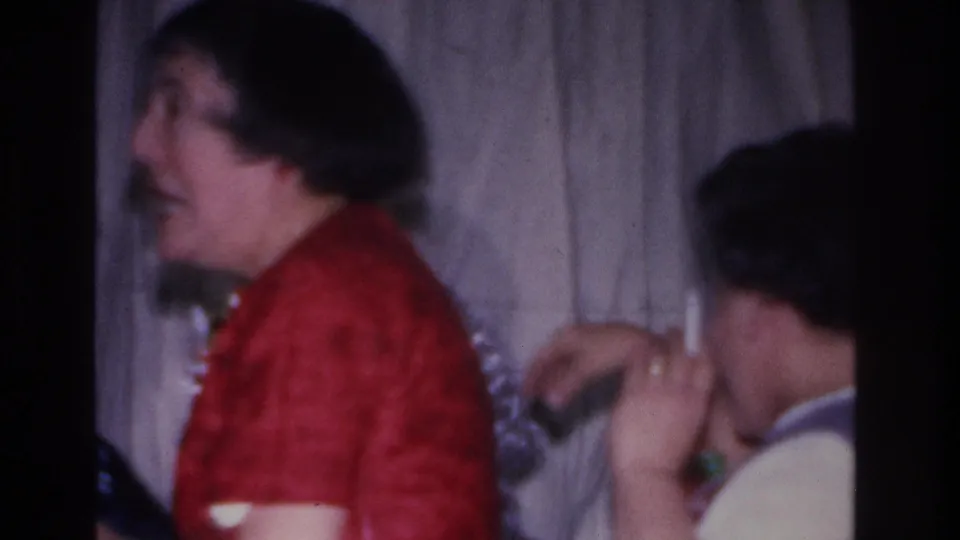Woman in red top in close profile, another woman behind her, curtain backdrop, 8mm home movie, mid-1960s indoor gathering