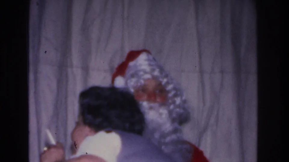 Santa Claus in red hat and white beard with woman leaning toward him in profile, curtain backdrop, 8mm home movie, mid-1960s holiday