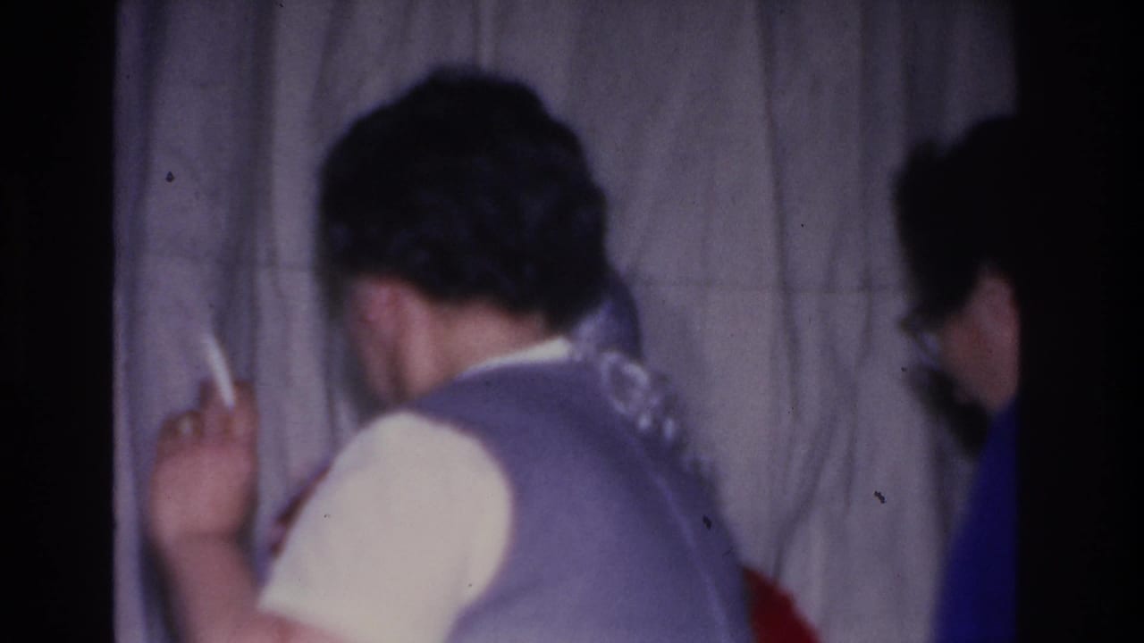 Two adults photographed from behind at a Christmas gathering in Hoboken, New Jersey, circa 1966; one in a blue-purple vest appears to hold a cigarette; gray curtain backdrop.