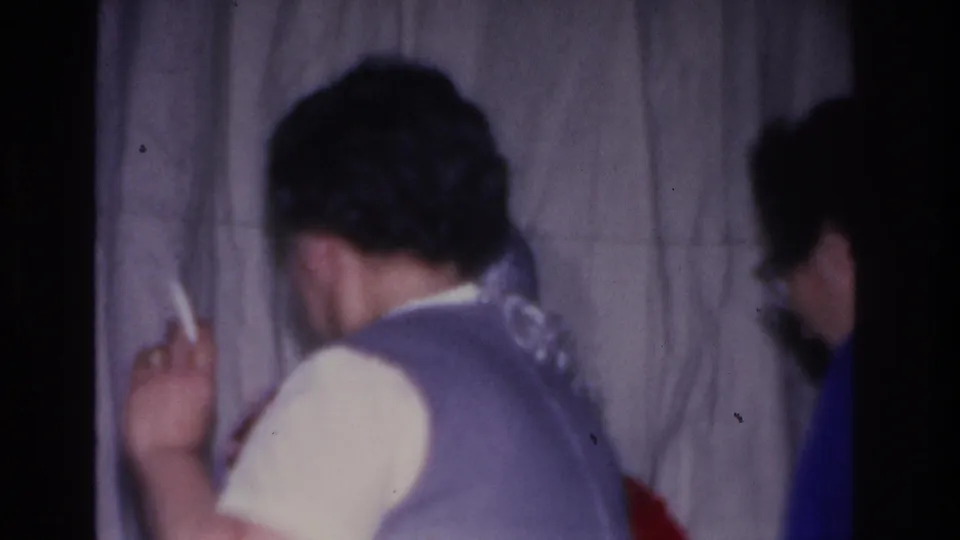 Two adults photographed from behind at a Christmas gathering in Hoboken, New Jersey, circa 1966; one in a blue-purple vest appears to hold a cigarette; gray curtain backdrop.