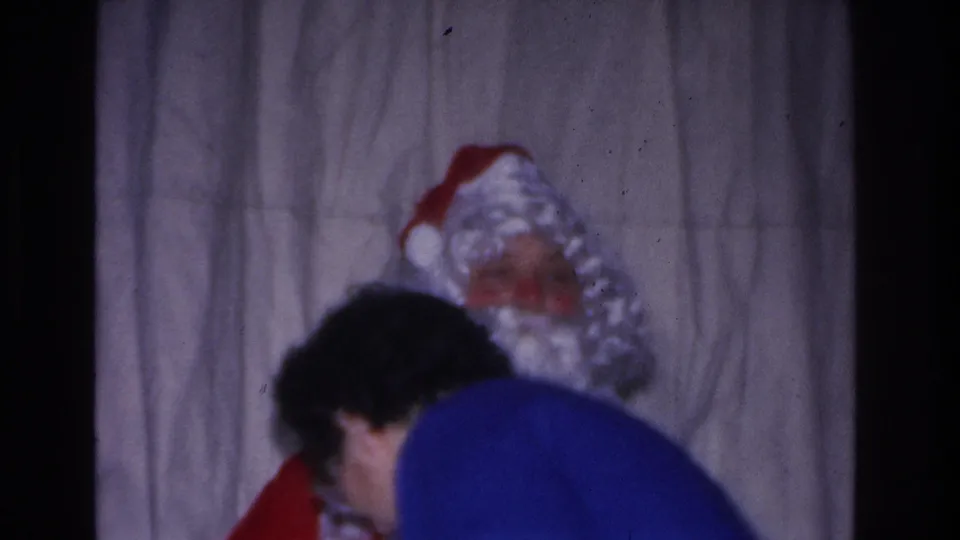 Santa Claus figure in red hat and white curly beard interacts with an adult in a bright blue top at a 1966 Christmas gathering in Hoboken, New Jersey; gray curtain backdrop.