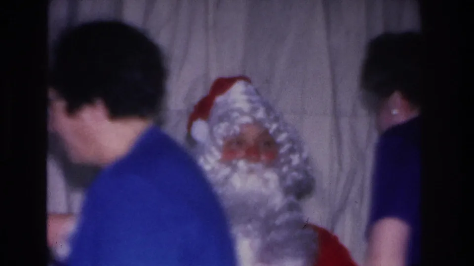 Santa Claus figure with two adults at a 1966 Christmas party in Hoboken, New Jersey; person in blue prominent in foreground, second adult visible in background; gray curtain.