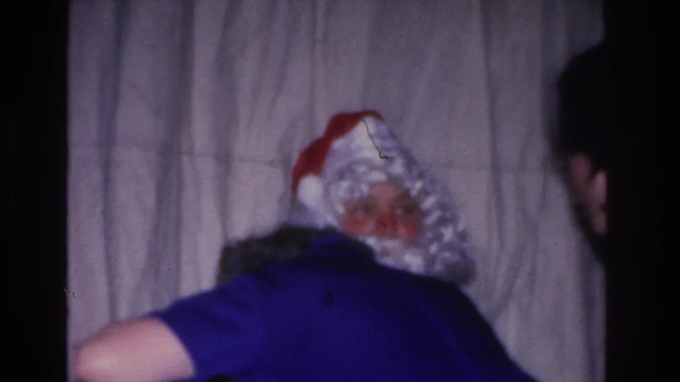 Motion captured as a guest in blue leans forward beside Santa Claus near the end of a 1966 Christmas gathering in Hoboken, New Jersey; third adult at right edge; gray curtain.