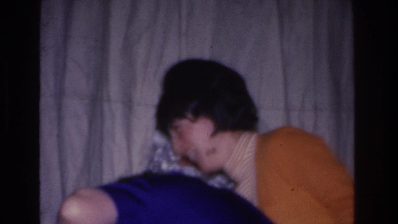 Person in a blue top and another in an orange sweater in close-up against a plain wrinkled curtain backdrop.
