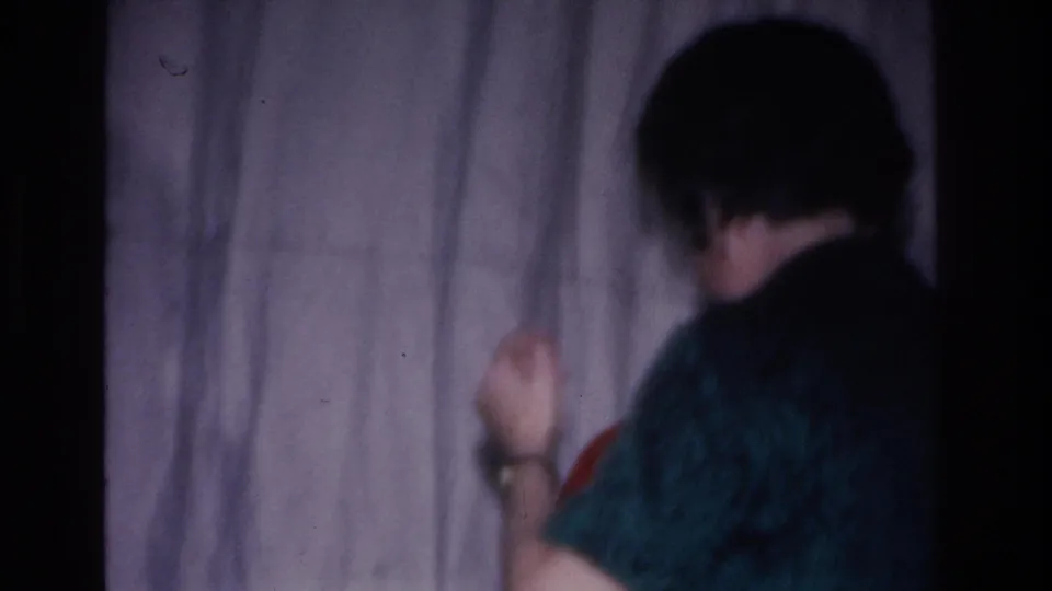 8mm home movie frame, c.1966: rear view of person in dark teal top with arm raised and wristwatch visible; Santa's red hat partially at lower left; plain curtain backdrop.