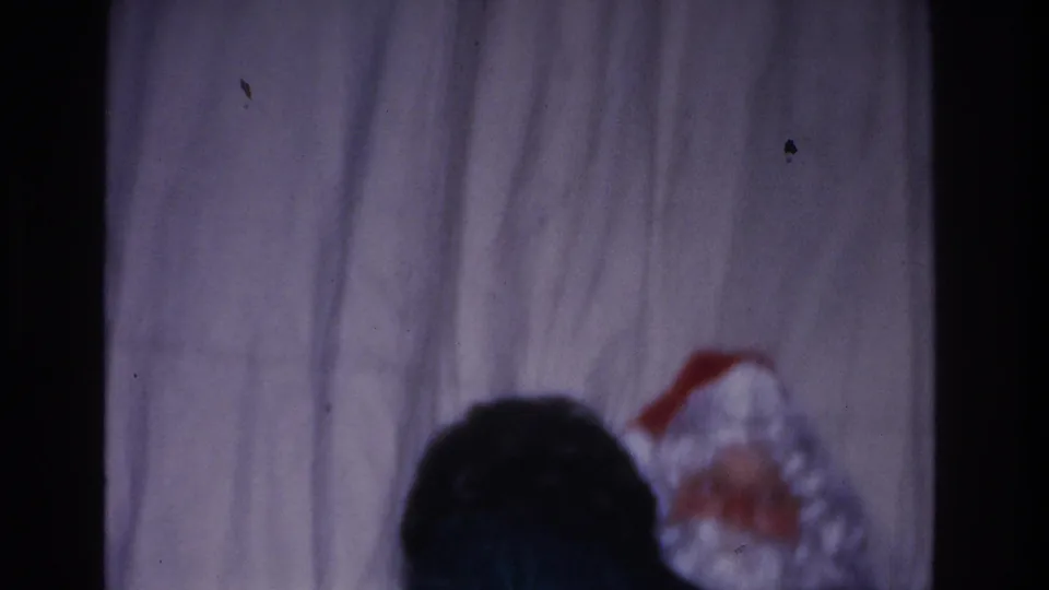 8mm home movie frame, c.1966, Hoboken NJ: dark-haired figure from behind in foreground; Santa Claus in red hat and white beard at lower right against a plain curtain.