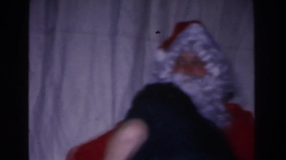 Santa Claus leans over a dark-clothed figure at what appears to be a 1966 Hoboken, New Jersey Christmas gathering, filmed on 8mm.