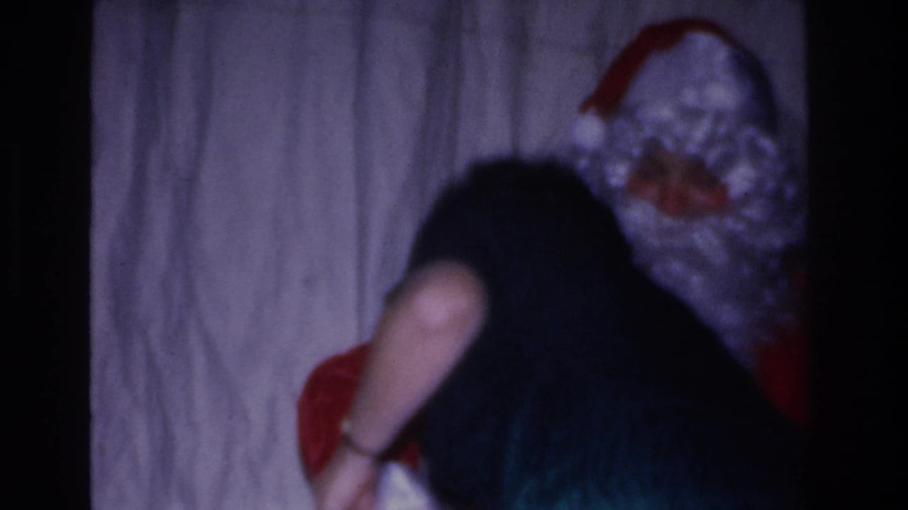 Santa costume figure bends toward a dark-haired person, with red suit fabric visible, at an indoor 1960s holiday gathering shot on 8mm.