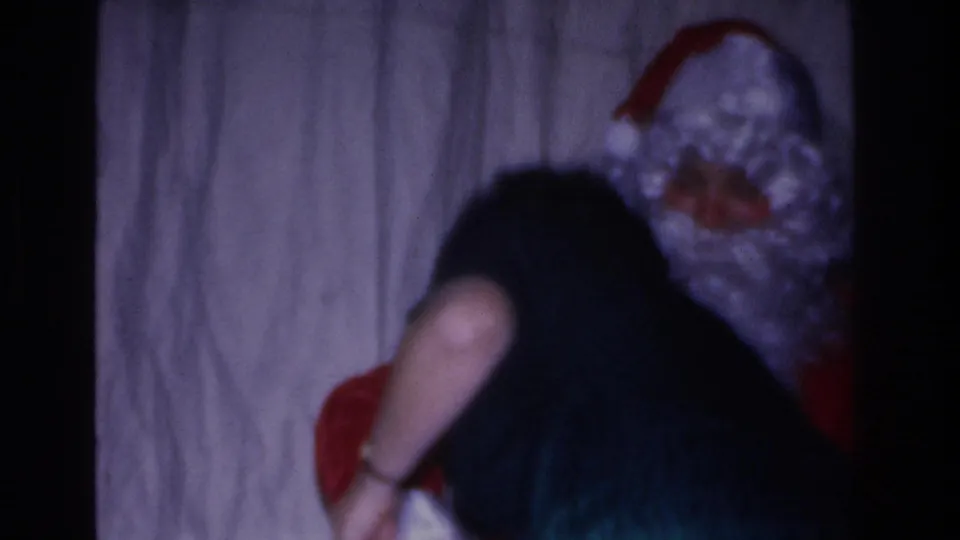Santa costume figure bends toward a dark-haired person, with red suit fabric visible, at an indoor 1960s holiday gathering shot on 8mm.