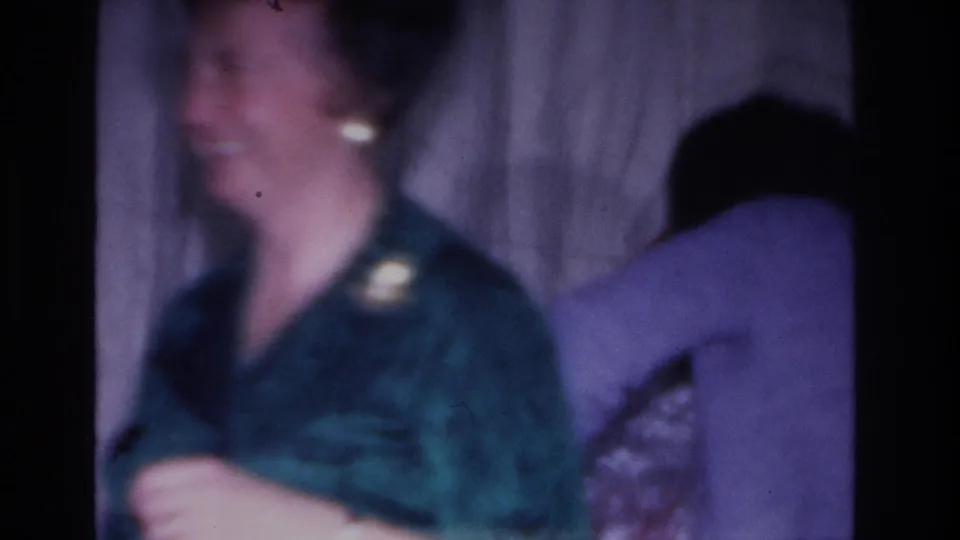 A smiling woman in a teal dress with a brooch and earrings reacts warmly at what appears to be a 1966 Hoboken holiday gathering; a second figure in lavender is partially visible behind her.