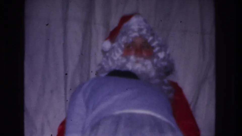 Rear view of a Santa Claus costume — red suit with white wig trim — bending forward at an indoor 1960s holiday gathering filmed on 8mm.