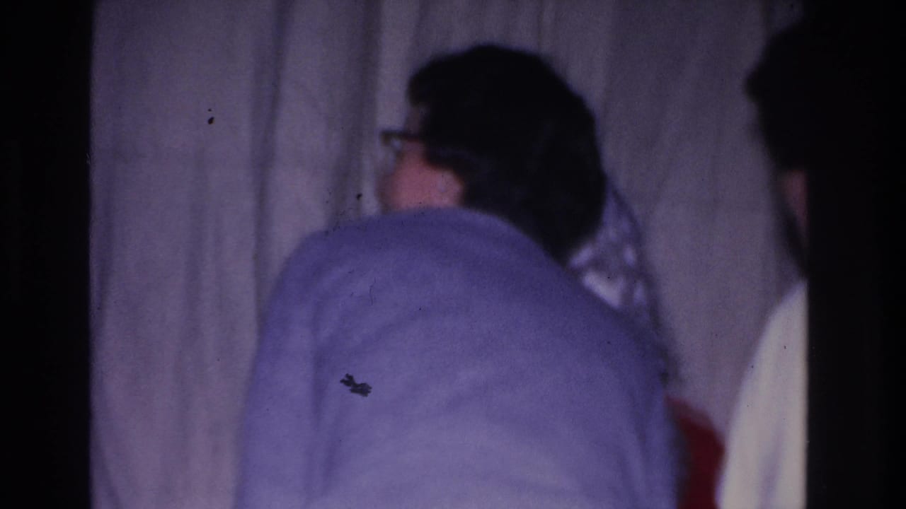 Adult in light-blue wool sweater seen from behind at a 1966 Hoboken, New Jersey community Santa visit; white curtain backdrop, 8mm purple color cast.