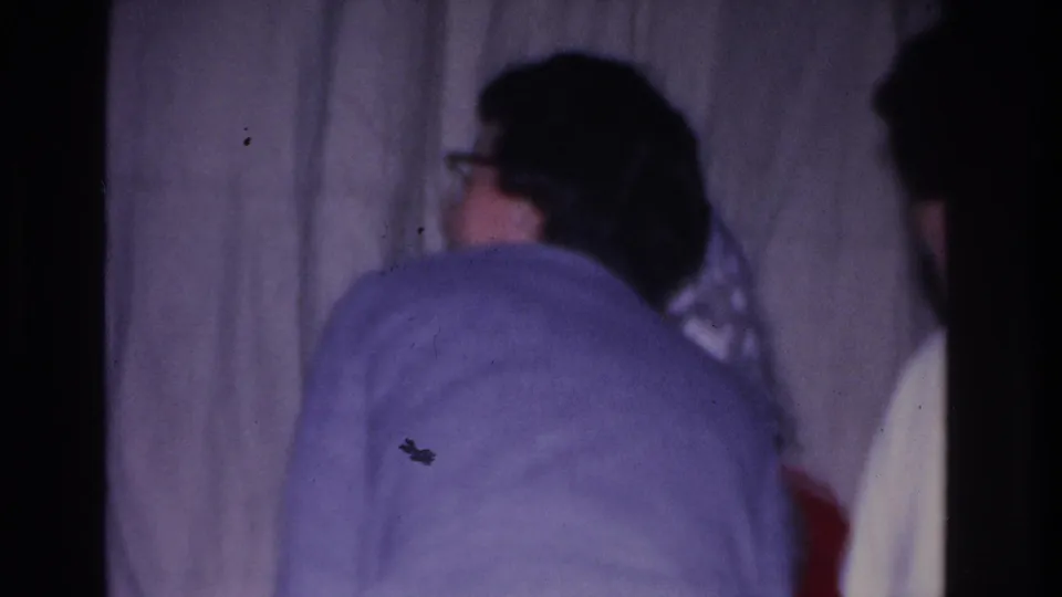 Adult in light-blue wool sweater seen from behind at a 1966 Hoboken, New Jersey community Santa visit; white curtain backdrop, 8mm purple color cast.