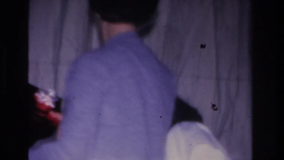 Adult in light-blue sweater filmed from behind at a 1966 Hoboken, NJ Santa visit; Santa's red costume partially visible at right, white curtain backdrop, 8mm film grain.