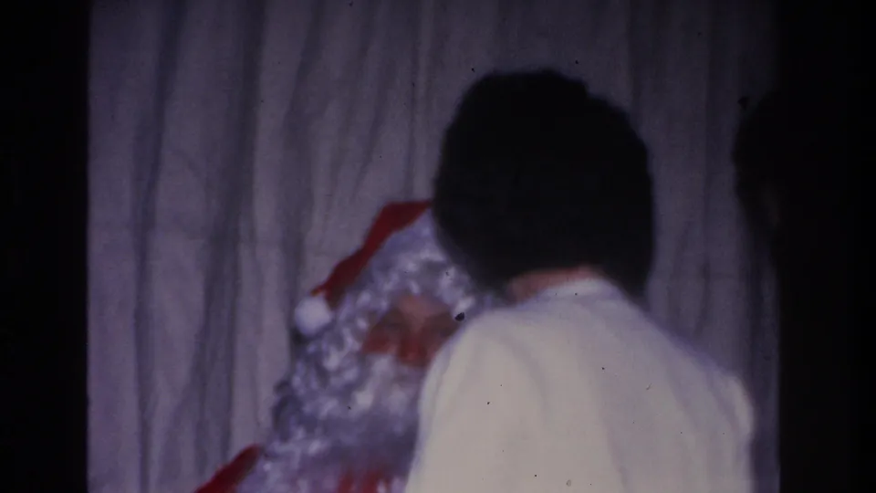 Person in white shirt leans toward Santa Claus — white curly wig and red suit visible — at a 1966 Hoboken, NJ community holiday event; draped curtain backdrop, 8mm film.