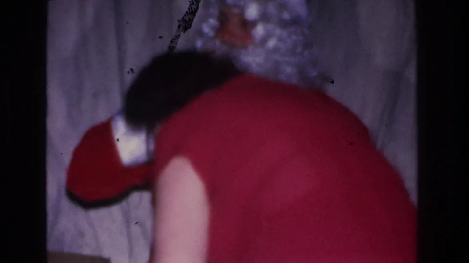 Blurred 8mm close-up, c. 1966; Santa's red suit with white and black trim seen from behind, woman's dark hair in the foreground at an indoor Christmas gathering.