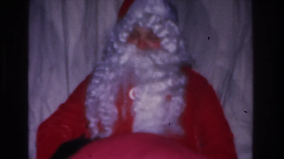 8mm home movie, c. 1966; rear view of Santa Claus in red suit, white curly wig and beard, and white gloves against a gray fabric curtain at an indoor Christmas event.