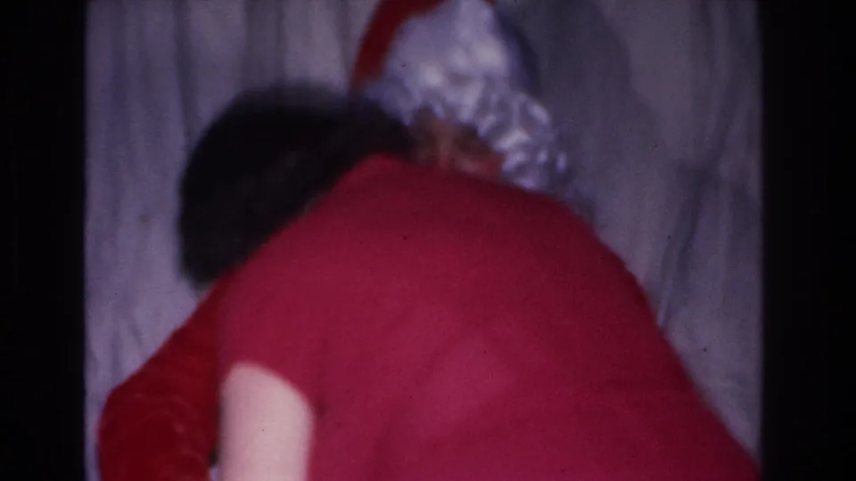 Woman in red dress leans toward a Santa Claus figure in a domestic interior; 8mm home movie, Hoboken, New Jersey, circa 1966.