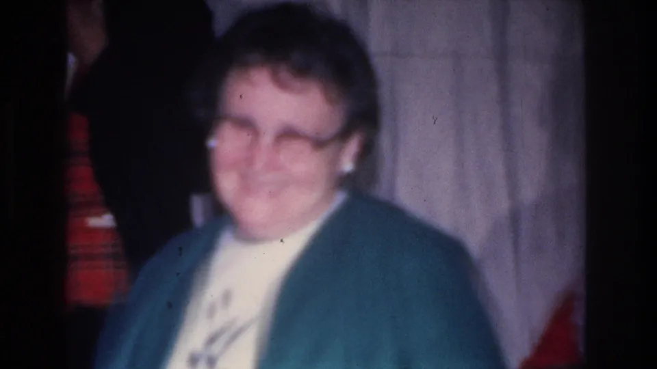 Close-up of a woman in glasses and teal cardigan over a white blouse, in a domestic holiday setting; 8mm footage, Hoboken, New Jersey, circa 1966.