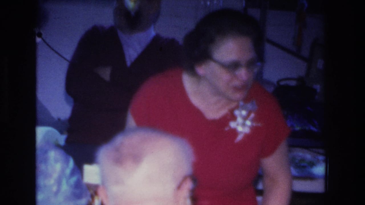 Woman in red dress wearing eyeglasses and white corsage at what appears to be a mid-1960s indoor social gathering filmed on 8mm in Hoboken, New Jersey.