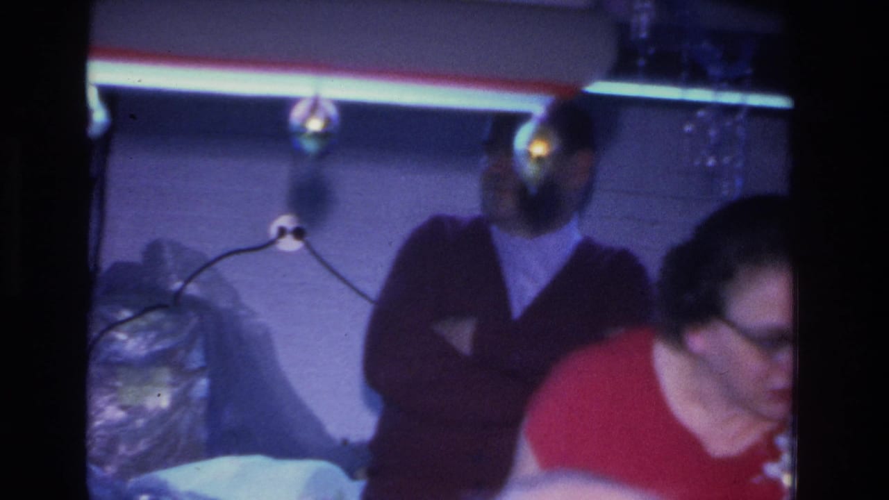 Man in dark sweater standing with arms crossed at what appears to be a mid-1960s indoor party; decorative bulbs and plastic-wrapped items visible in background.