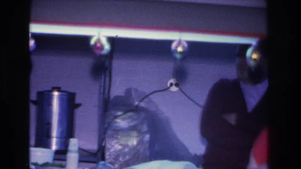 Refreshment table with a large silver electric percolator and covered items at a mid-1960s American indoor gathering; decorative bulbs hang above the table.