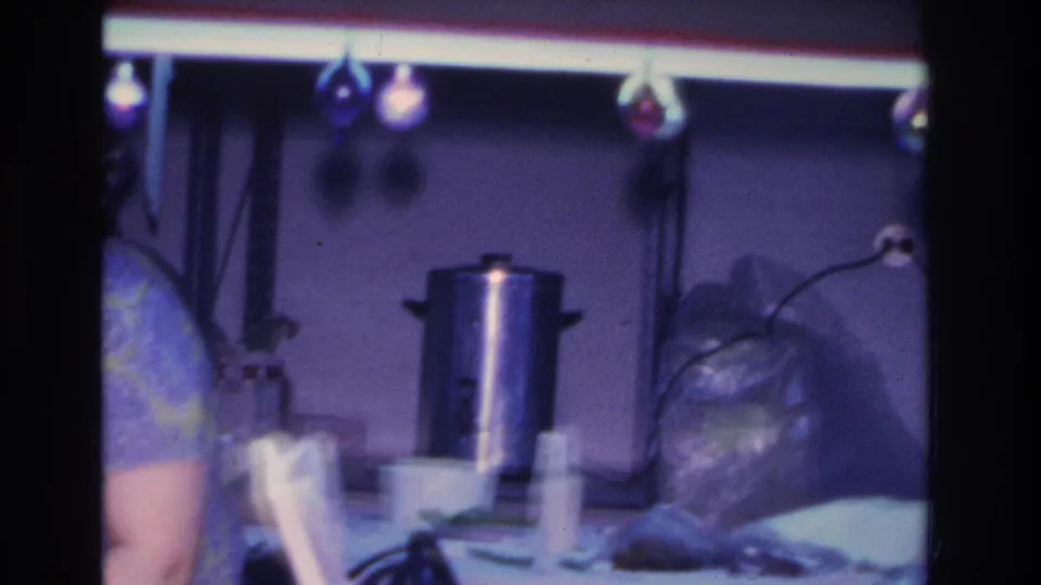 Refreshment area at a mid-1960s indoor gathering showing a large silver coffee urn, covered items on the table, and overhead decorative lights.