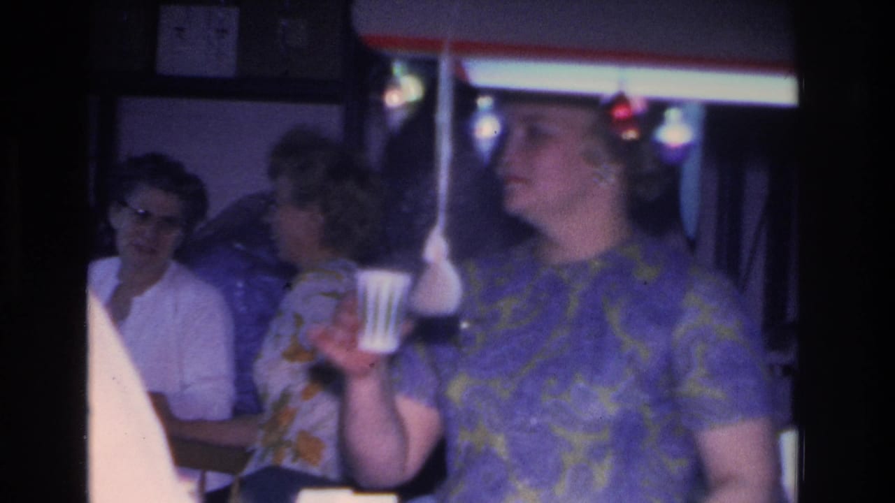 Woman in patterned dress holds a white cup; two other women socialize behind her beneath decorative overhead lights.
