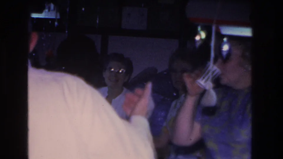 A crowded party interior with a woman wearing glasses visible mid-frame smiling, another figure raising a cup to drink, and colorful string lights above, from a 1966 8mm home movie shot in Hoboken, New Jersey.